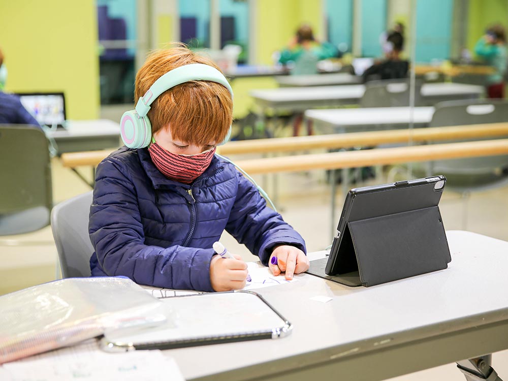 Young boy working on school with a mask and headphones on.